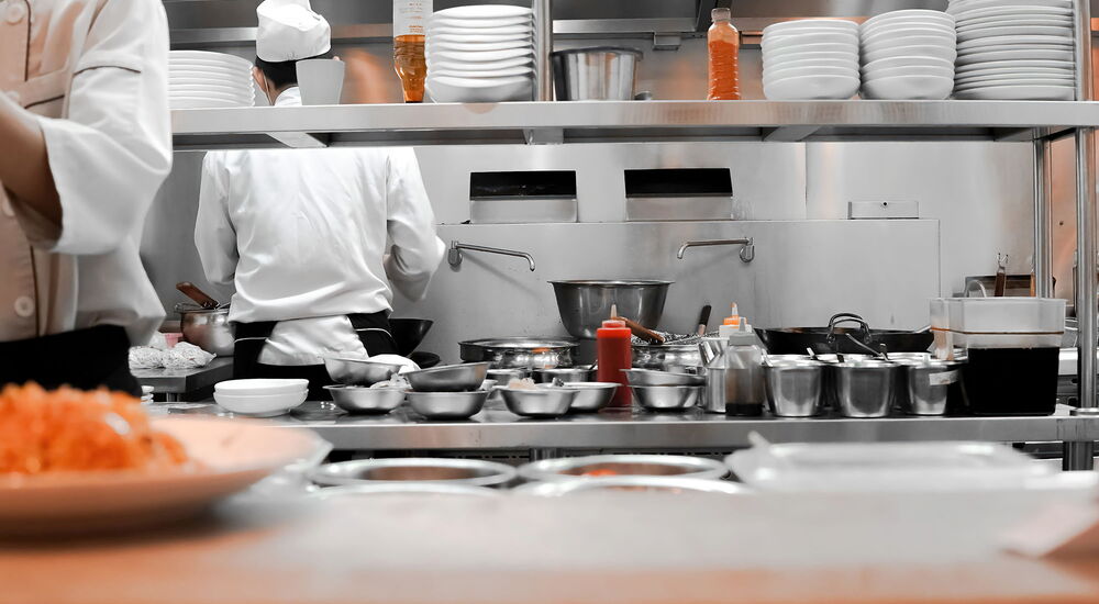 Eine Küche mit vielen Töpfen und Tellern. Ein Koch ist von hinten zu sehen, der andere nur angeschnitten vorn vorn.