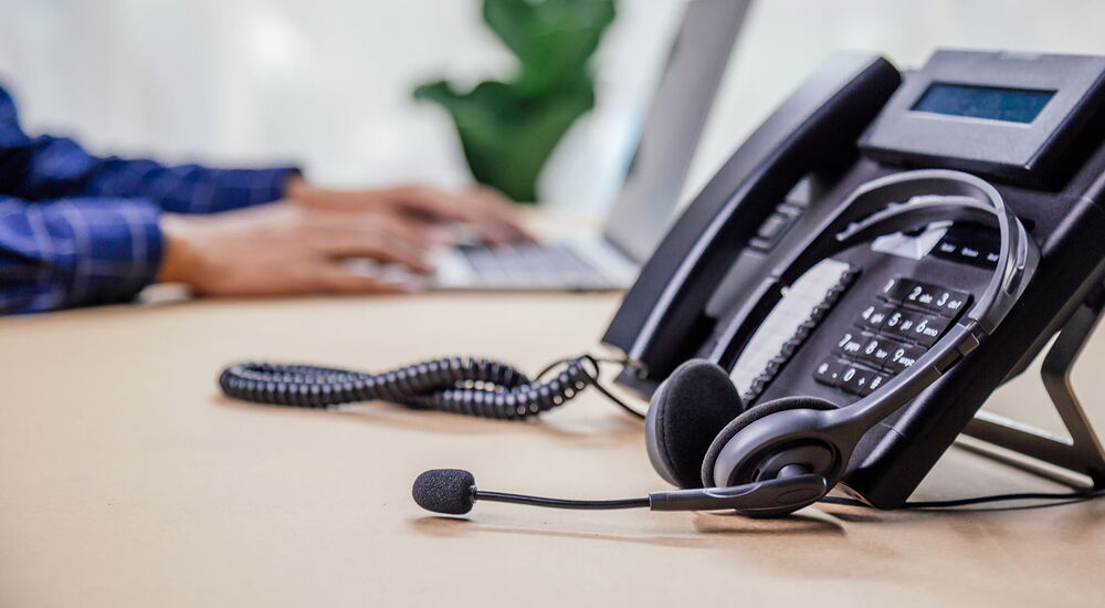 Ein Telefon mit Headset steht auf einem Schreibtisch - im Hintergrund sind Hände auf einer Laptoptastatur zu sehen.