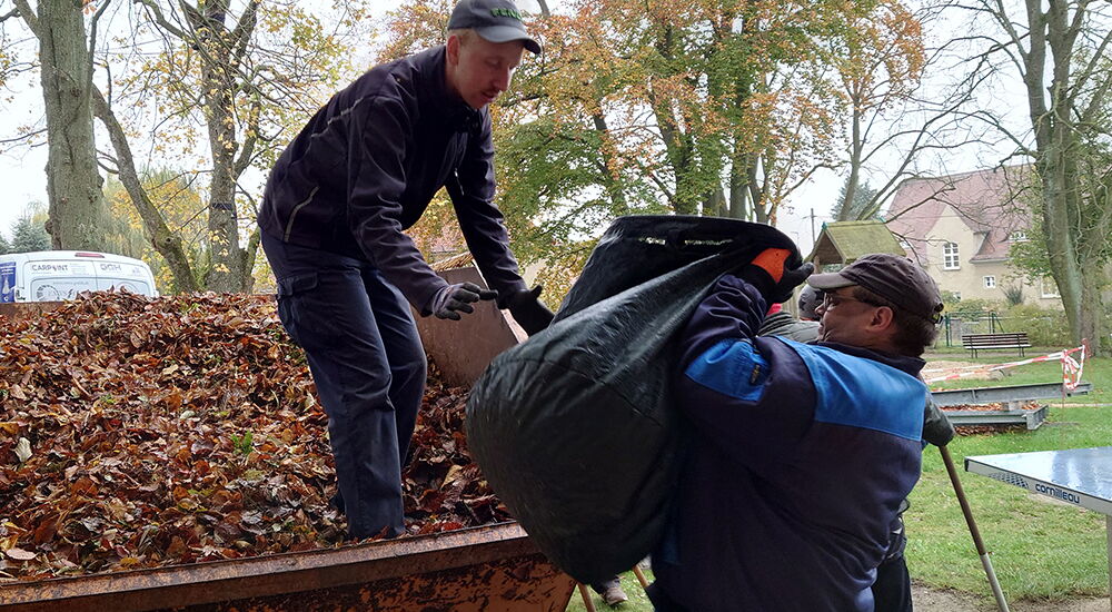Zwei Werkstattbesucher heben einen großen Sack mit Blätter in einen Container.